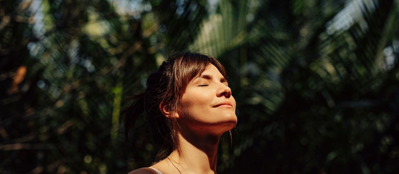 A woman standing outdoors with her eyes closed, face turned towards the warm afternoon light